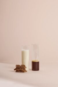 Transparent glass flasks filled with milk and cocoa placed near chocolate near light pink background